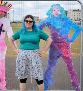 young woman with blue hair stands with her hands on her hips, mirroring the pose of the paper silhouette of herself next to her, thats painted in bright blue, pink and purple