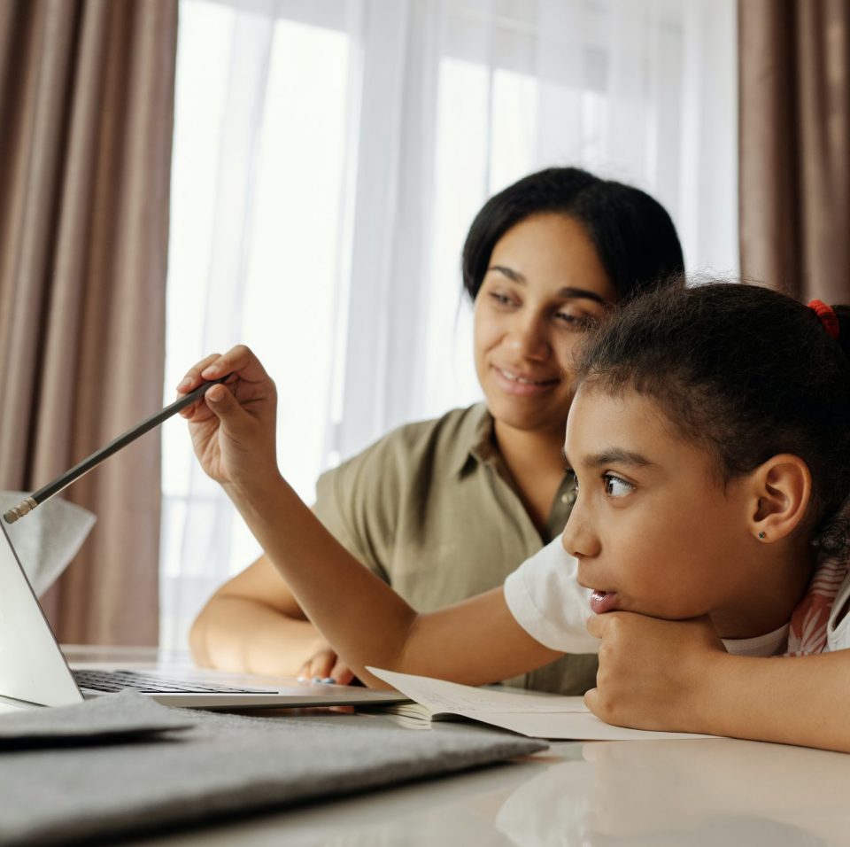 Brown woman and pre-teen girl sit together at a table, the girl gesturing with a pen