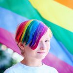 Child with hair coloured into a rainbow, standing in front of a rainbow flag