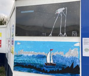 Photo of two large painted artworks, the lower one a seascape in many shades of blue with thick expressive brush stroke, a light blue sky behind, and a yacht toward the centre of the image. The upper artwork is almost totally black, with light dots, possibly stars. The focus of the piece is something with three long thing straight legs, each splitting into three at the bottom. They connect with some kind of body at the top, bent over and pointed at one end.