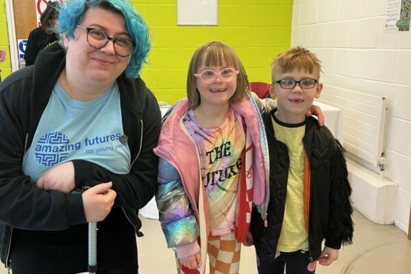 Amazing Futures volunteer Eliph, a White young woman with blue hair, leans on a walking stick as she leans down to pose with two children in glasses, all smiling at the camera.