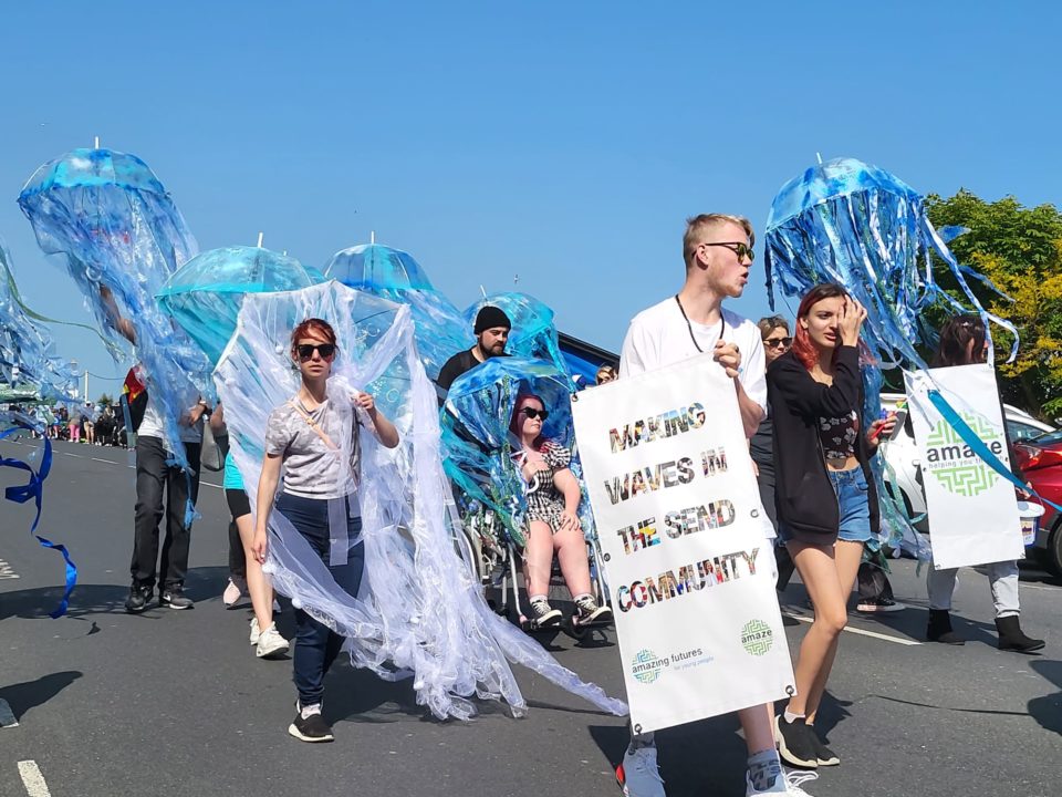 Young people in various jellyfish costumes, one using a wheelchair, some holding signs, a crowd is visible behind at the road edge.