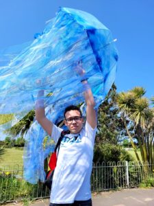 Young man holding jellyfish umbrella sideways high in the air, the tendrils floating behind it in the wind, and a bright blue sky is behind them.