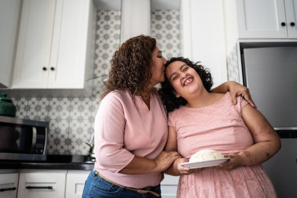 Young learning disabled woman and her mum laugh and cuddle in a kitchen.