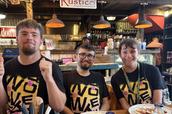 Three young people in matching t-shirts at a pizza restaurant, putting toppings on pizza