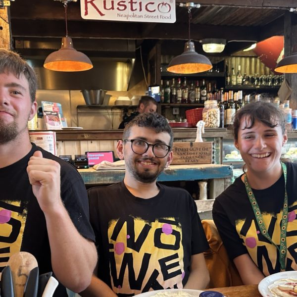 Three young people in matching t-shirts at a pizza restaurant, putting toppings on pizza